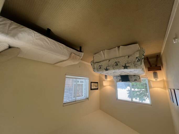 Your cozy loft bedroom features a unique bunk bed tucked beneath soaring ceilings, two bright windows for natural light, and soft neutral tones creating a peaceful retreat.