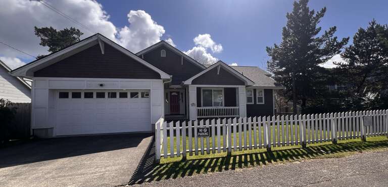 Charming cottage-style vacation rental featuring a white picket fence, attached garage, and covered front porch in a peaceful residential neighborhood surrounded by evergreen trees.