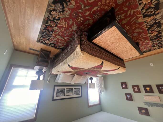 Your cozy bedroom awaits with a plush bed and ornate decorative ceiling. Warm wood furnishings, natural light from the windows, and framed art create a welcoming retreat where you can unwind.