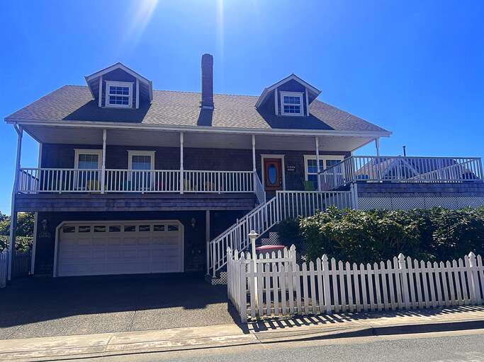 Charming coastal property featuring classic architecture with spacious wraparound porches, dormered rooflines, and a convenient ground-level garage, framed by mature hedges along a quiet street.