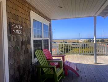 Seaview Manor welcomes you with a covered deck featuring colorful Adirondack chairs and ocean views stretching across the dunes and sparkling blue water beyond.