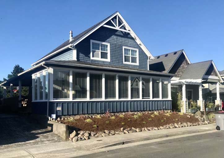 A charming Craftsman-style home with classic dark blue siding and white trim greets visitors from a quiet residential street. The terraced stone landscaping and blooming garden beds frame the welcoming front porch.