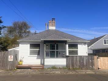 A charming single-story cottage with a hip roof and brick chimney, featuring a welcoming front porch and classic white siding in a quiet residential neighborhood.