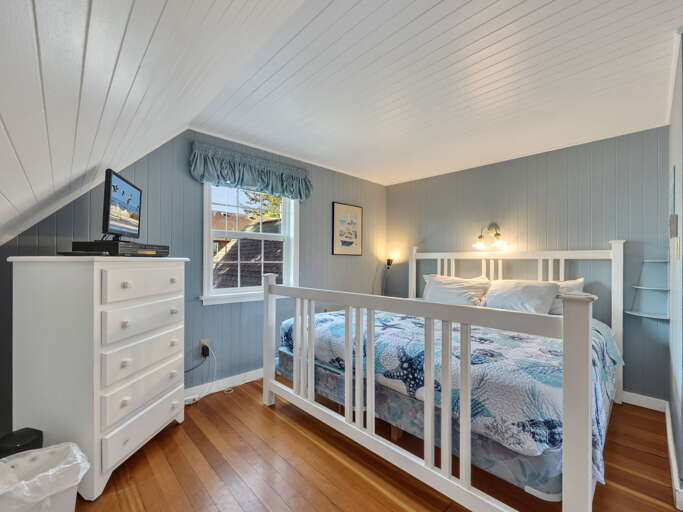 Cozy loft bedroom with warm wood floors and charming coastal colors. Your comfortable bed awaits under vaulted ceilings, with soft lighting and a dresser for all your belongings.