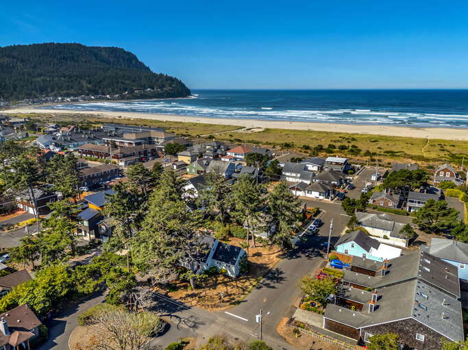 Aerial view of a charming coastal neighborhood nestled between forested hills and a pristine sandy beach, with ocean waves rolling in and scenic natural surroundings.