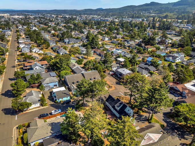 Aerial perspective of a charming coastal community nestled between tree-lined streets and rolling hills, offering a peaceful neighborhood setting with ocean glimpses in the distance.