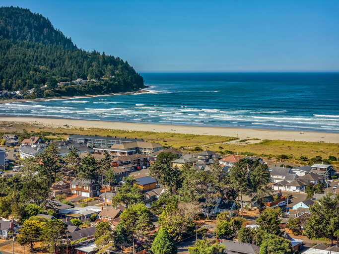 A charming coastal community nestled between forested mountains and pristine sandy shores, with the sparkling Pacific stretching to the horizon beyond.