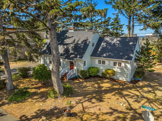 Charming coastal home nestled among towering evergreens, with ocean glimpses visible through the trees. The property features mature landscaping and a peaceful, forested setting near the water.