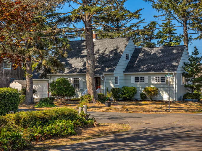 A charming light blue cottage nestled among mature trees on a quiet residential street, offering a peaceful retreat surrounded by greenery and natural shade.