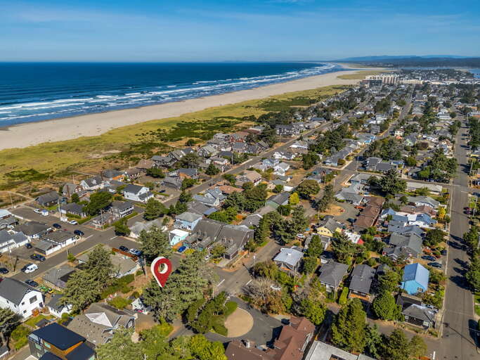 Aerial view of a coastal neighborhood situated along a stunning stretch of sandy beach. The property is located within a residential area surrounded by mature trees, with easy access to both the waterfront and local amenities.