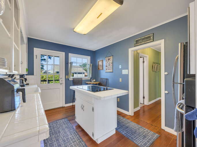 Step into your bright kitchen where ocean breezes flow through charming French doors, warm wood floors welcome your bare feet, and a central island invites you to gather for morning coffee or meal prep with garden views just beyond.