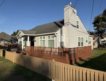 A charming coastal cottage with light-toned siding and expansive deck welcomes guests in a quiet, tree-lined neighborhood under a clear blue sky—the perfect retreat for your getaway.