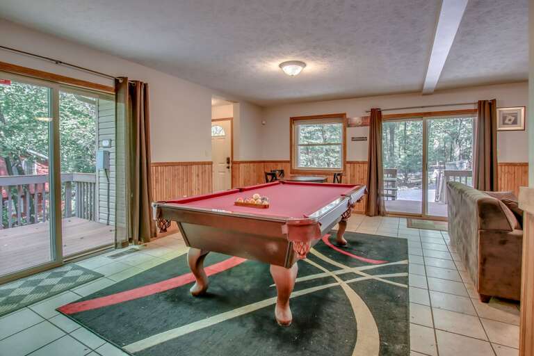 A pool table in the middle of the game room on a multicolored rug, with a couch to the right and sliding glass door to the left and behind. A pool table in the middle of the game room on a multicolored rug, with a couch to the right and sliding glass door to the left and behind.