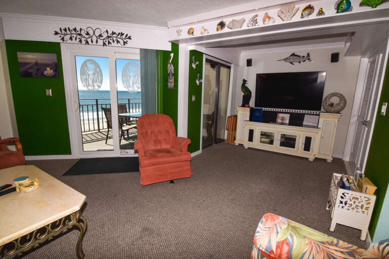 Seaside Stay Visible Through Balcony Doors, Colorful Chairs Complement Carpeted Living Area