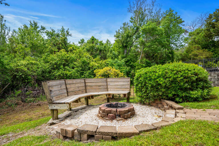 Backyard Bench Curving Near A Bricked Fire Pit Surrounded By Bushy Greenery And A Bright Blue Sky