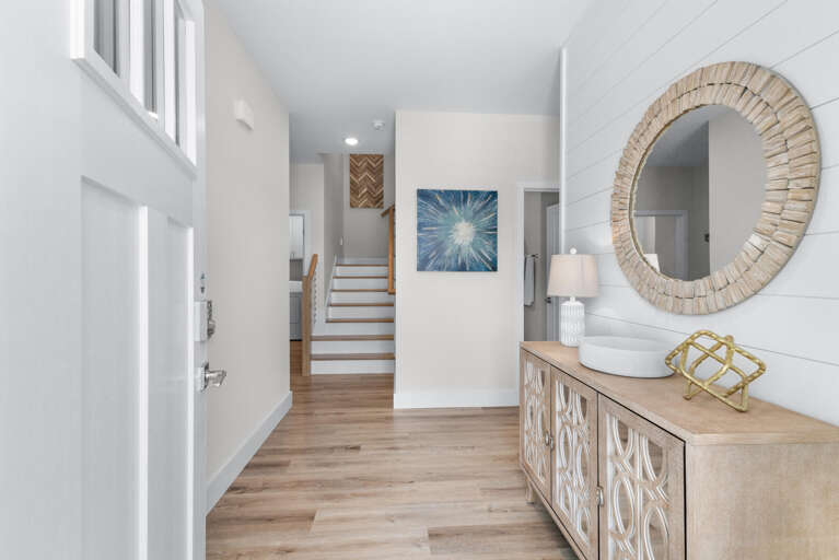 Bright, Beachy Vacation Rental Entryway With White Walls, Wooden Floor, Round Mirror, And Cabinet