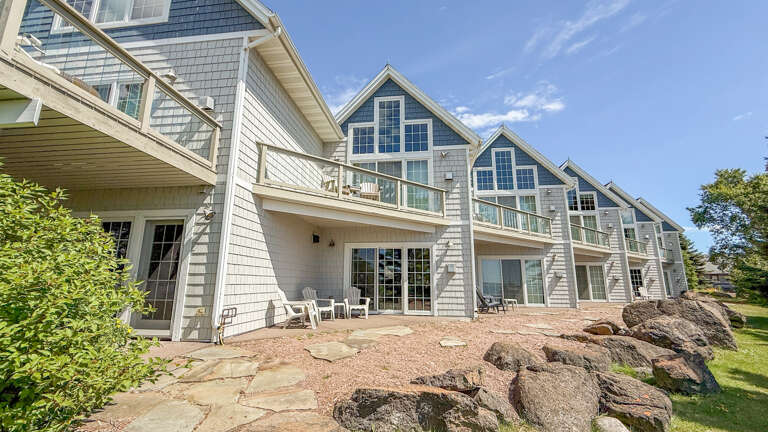 This charming shingled property features multiple units with private patios and balconies, all nestled among natural stone landscaping and lush greenery under a bright blue sky.