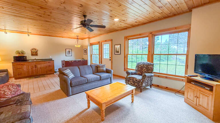 Unwind in your spacious living area with warm wood ceilings, cozy seating, and bright windows framing peaceful garden views—your perfect retreat after a day of adventure.
