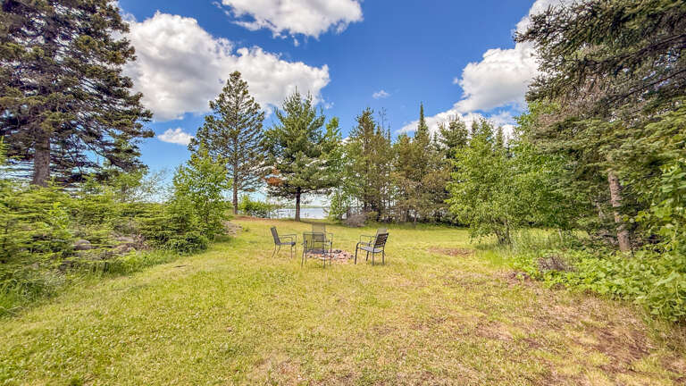 Peaceful natural setting featuring a fire pit surrounded by seating, framed by towering pines and open sky—a serene spot on the property grounds.