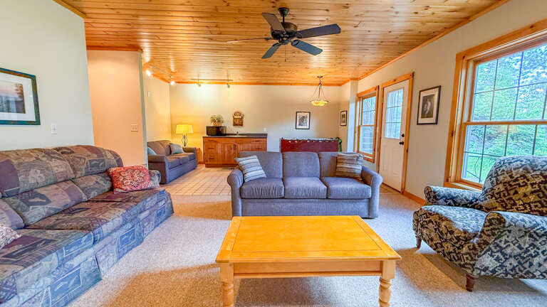 Unwind in your spacious living room with vaulted pine ceilings, plush seating for the whole group, and natural light streaming through large windows framing peaceful wooded views.