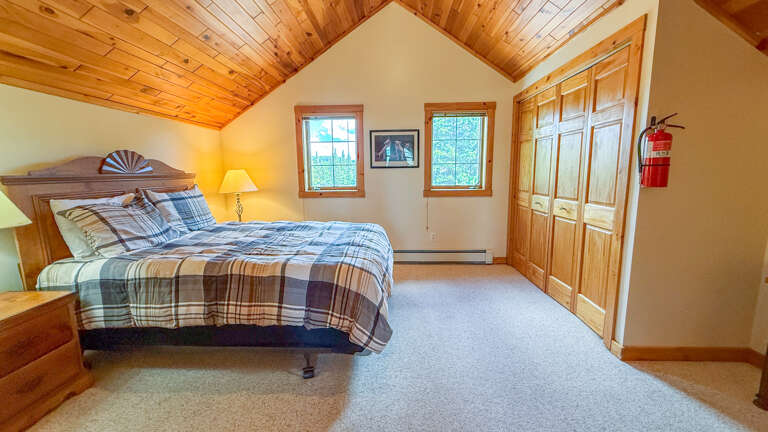 Drift off under warm wood-paneled ceilings in this cozy bedroom, where natural light pours through double windows and plush plaid bedding invites your best night's sleep.
