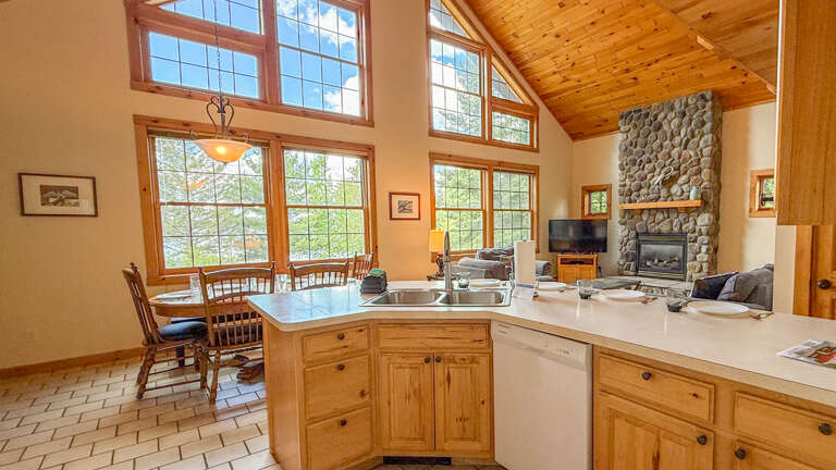 Sunlight floods this open kitchen and living space, where towering windows frame forest views, a stone fireplace anchors cozy moments, and vaulted pine ceilings create a warm, airy mountain retreat.