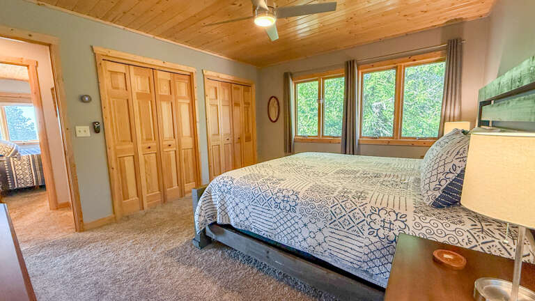 Your peaceful bedroom retreat features a plush bed beneath a stunning pine-planked ceiling, with large windows framing lush greenery that fills the space with natural light and tranquility.
