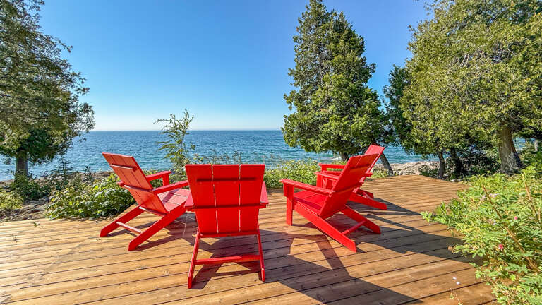 This waterfront deck offers stunning views where vibrant red Adirondack chairs frame a tranquil lakeside setting, framed by towering trees and natural shoreline.