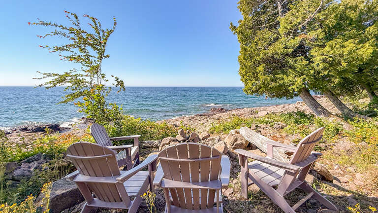This waterfront retreat offers direct shoreline access where guests can relax in Adirondack chairs surrounded by natural lakeside beauty and mature trees.