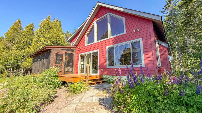 A cheerful red cottage with large windows and a wood-screened patio, nestled among towering trees and vibrant purple wildflowers—your peaceful escape awaits.