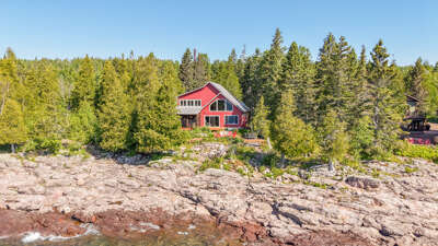 A charming red cabin nestled among evergreens along a rocky shoreline, offering peaceful waterfront seclusion and natural beauty.
