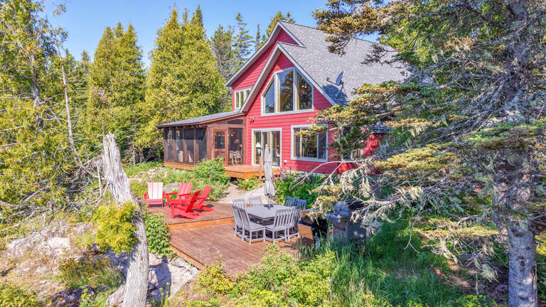 A charming red cottage nestled in lush forest surroundings with a spacious wooden deck and outdoor dining area, perfect for enjoying nature during your stay.