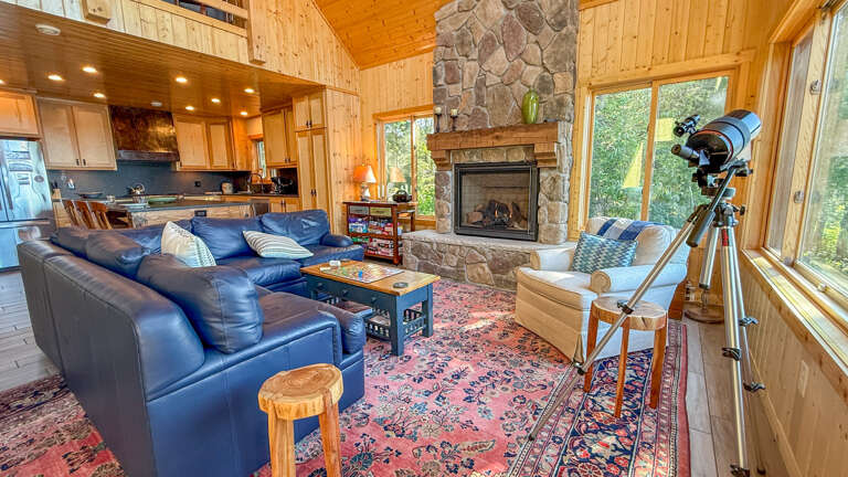 Relax in your inviting living room featuring a stone fireplace, cozy leather seating, and stunning forest views through expansive windowsâperfect for unwinding after a day of adventure.