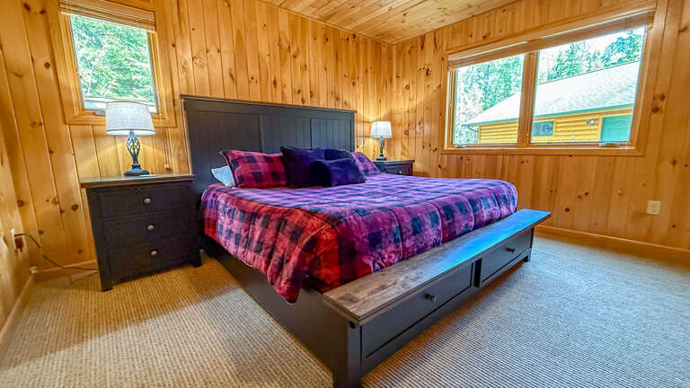 Your cozy cabin bedroom invites restful sleep with its warm wood walls and plush plaid bedding. Wake to natural light streaming through large windows overlooking peaceful evergreens.
