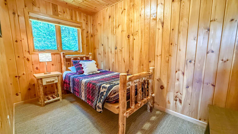 Cozy up in this warm wooden bedroom with its rustic log-style bed and inviting quilted bedding. Natural light filters through the window, offering a peaceful view of lush greenery.
