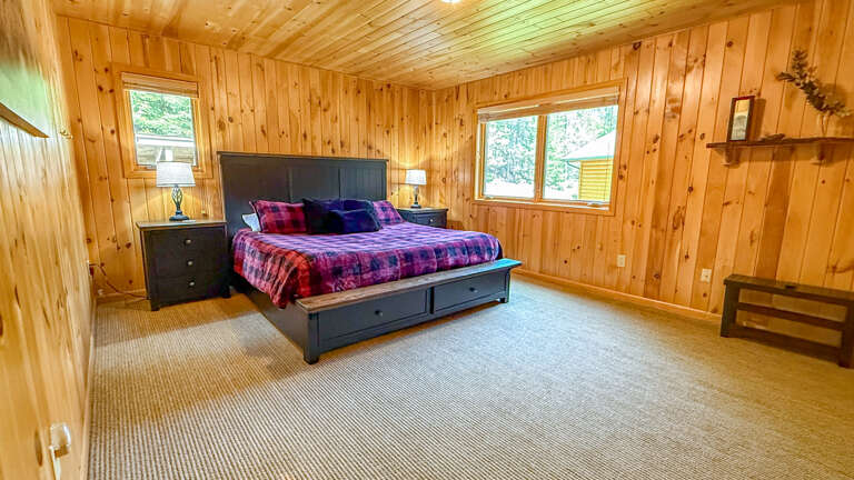 Relax in your spacious bedroom with rustic pine walls, a cozy king bed with plaid linens, and natural light streaming through windows overlooking greenery.