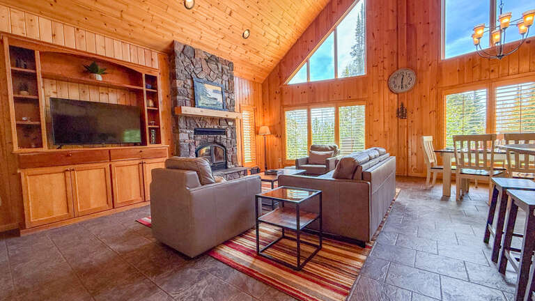 Settle into your cozy living space with vaulted ceilings, a stone fireplace, and plush seating surrounded by warm wood tones and natural light streaming through expansive windows.