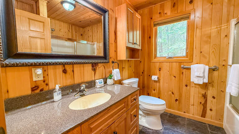 Step into your charming cabin bathroom, where warm knotty pine walls create a cozy, rustic retreat. Fresh towels await while natural light filters through the forest view.