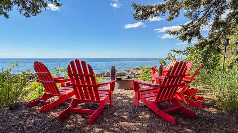 Crimson Adirondack chairs frame a fire pit overlooking the calm lakeâyour perfect spot to unwind with morning coffee or evening conversations by the shore.