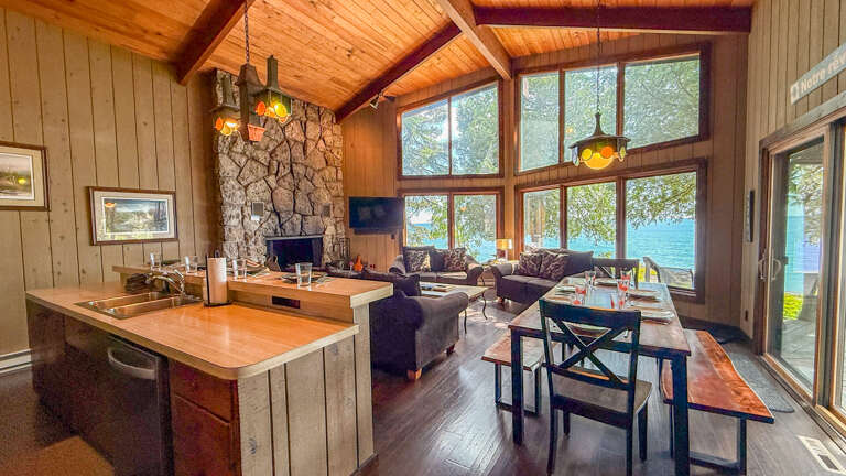 Gather around the handcrafted dining table or sink into the cozy seating area as natural light floods through floor-to-ceiling windows framing serene lake views. The stone fireplace and vaulted wood ceilings create warmth and rustic charm.