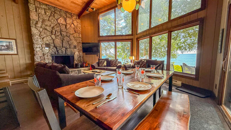 Gather around the stunning live-edge dining table as sunlight streams through floor-to-ceiling windows, framing breathtaking lake views. The cozy stone fireplace and vaulted wood ceilings create the perfect setting for memorable meals.