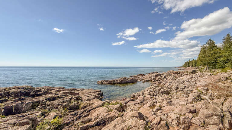 Rocky shoreline meets calm waters under a bright sky, surrounded by natural landscape and seasonal greenery in the area.