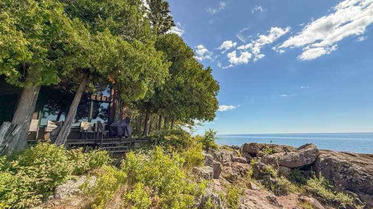 Waterfront property nestled among towering trees along a pristine rocky shoreline, with open sky and tranquil blue waters stretching to the horizon.