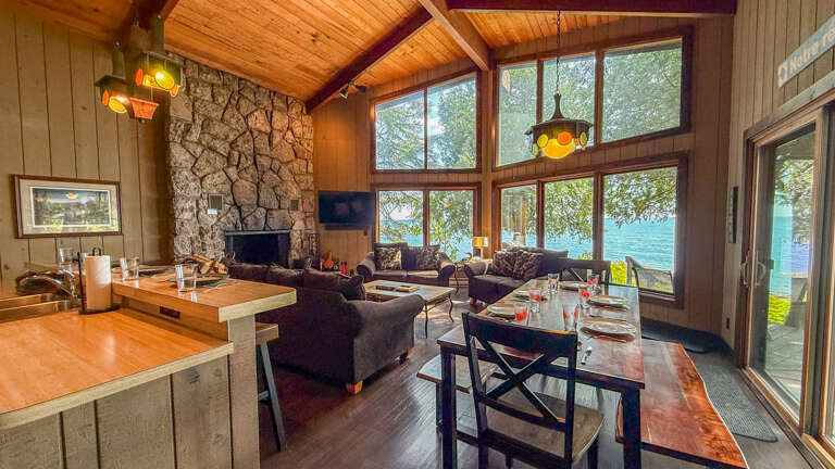 Unwind in this stunning open-concept space with soaring wood-beamed ceilings and floor-to-ceiling windows framing breathtaking lake views. Gather around the cozy stone fireplace or enjoy meals at the inviting dining area.