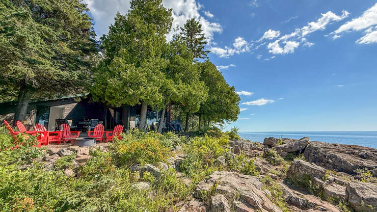Waterfront property nestled among towering pines on a rocky shoreline with bright red Adirondack chairs and expansive lake views.