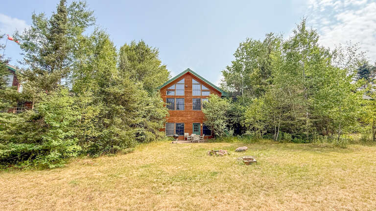 A charming wooden cabin nestled among tall trees with a spacious lawn, perfect for relaxing outdoors or gathering around the firepit under open skies.