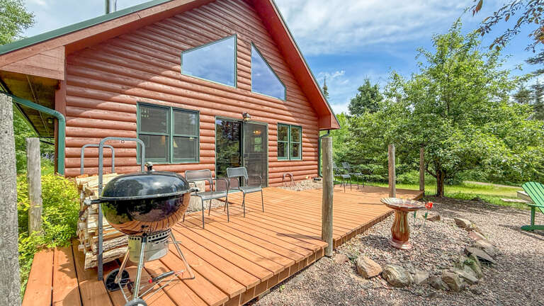 Charming log cabin exterior with spacious wooden deck, BBQ facilities, and outdoor seating surrounded by mature trees and natural landscaping.