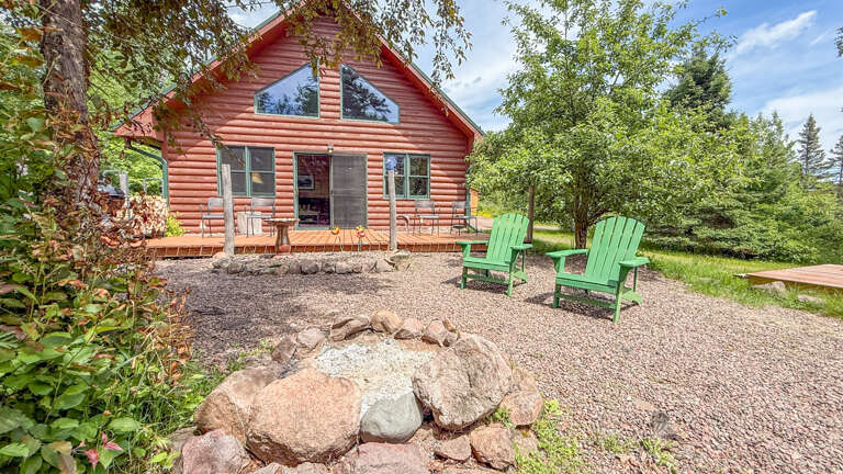 A charming red log cabin nestled among lush greenery, offering a peaceful retreat with a welcoming deck, fire pit, and Adirondack chairs for relaxing outdoor moments.