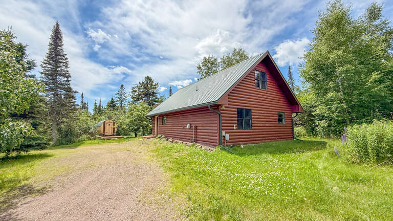 Charming red log cabin nestled among towering evergreens and lush greeneryâan inviting arrival at your private woodland retreat.
