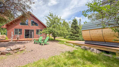 A charming red log cabin set amid mature trees and greenery with an outdoor sauna nearby for ultimate relaxation after exploring the natural surroundings.
